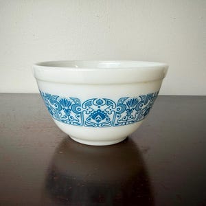 May include: White mixing bowl with a blue floral pattern around the exterior. The bowl is made of a thick, opaque material and sits on a dark wooden surface. The blue design is symmetrical and detailed.