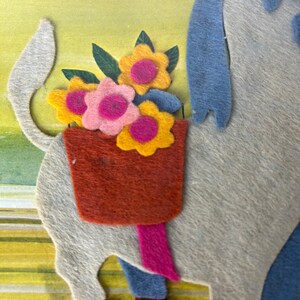 K&ouml;nnte beinhalten: Eine Filzapplikation eines wei&szlig;en Tieres mit blauem und grauem K&ouml;rper, das einen Korb mit gelben, rosa und orangefarbenen Blumen tr&auml;gt. Das Tier hat einen langen Schwanz und tr&auml;gt rote Schuhe.