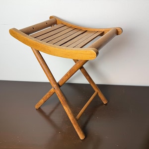 May include: A small, folding wooden stool with a light brown finish. The seat is made of parallel wooden slats. The legs are crossed and angled, providing stability. The stool is designed for portability and compact storage.