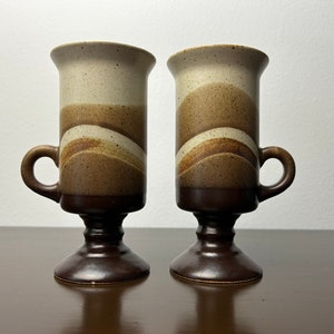 Puede incluir: Dos tazas de cerámica marrón y blanca con base de pedestal. Las tazas tienen un patrón de rayas marrón, blanco y beige.