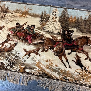May include: A tapestry depicting a winter scene with a horse-drawn carriage and a sled pulled by dogs. The scene includes figures in winter clothing, trees, and a snowy landscape. The tapestry has a beige fringe.