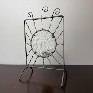 May include: A gray metal stand featuring a rectangular frame with wavy wire rays extending from a central circular glass insert. The glass displays a molded sun face design. The frame is topped with three decorative wire swirls.