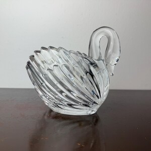 May include: A clear glass swan ornament with a curved neck and detailed feather design. The swan is on a dark brown surface. The ornament is a decorative item.