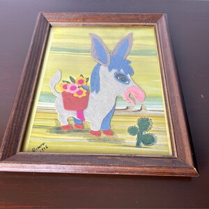 K&ouml;nnte beinhalten: Ein gerahmtes Filz-Applikationsbild eines Esels, der einen Korb mit Blumen tr&auml;gt. Der Esel ist grau mit blauen Ohren und einer rosa Nase. Der Hintergrund ist gelb mit gr&uuml;nen Streifen. Das Kunstwerk ist signiert mit "Dimitri 1116".