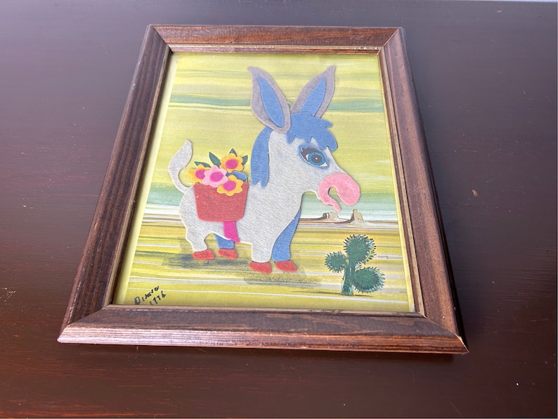 K&ouml;nnte beinhalten: Ein gerahmtes Filz-Applikationsbild eines Esels, der einen Korb mit Blumen tr&auml;gt. Der Esel ist grau mit blauen Ohren und einer rosa Nase. Der Hintergrund ist gelb mit gr&uuml;nen Streifen. Das Kunstwerk ist signiert mit "Dimitri 1116".