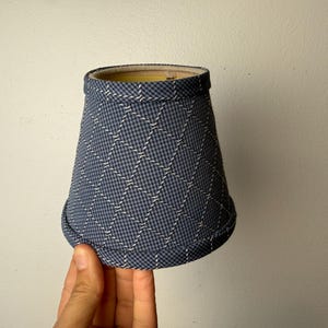May include: A blue and white patterned lampshade with a diamond grid design. The lampshade is held by a hand against a plain white background. The lampshade has a slightly flared shape.