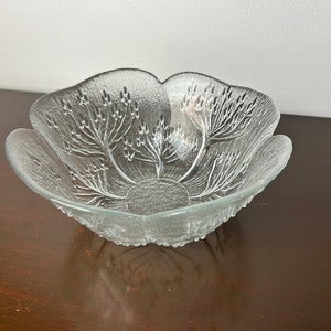Pode incluir: Uma tigela de vidro transparente com um design floral. A tigela tem uma borda festonada e uma superfície texturizada.