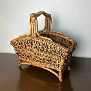 May include: A woven wicker basket with a handle. The basket is brown and has a natural finish. It is sitting on a dark brown surface.