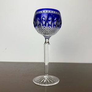 May include: A clear crystal wine glass with a cobalt blue bowl and a long, clear stem. The bowl features intricate cut patterns and a silver rim. The glass is designed for serving wine and adds a touch of elegance to any table setting.