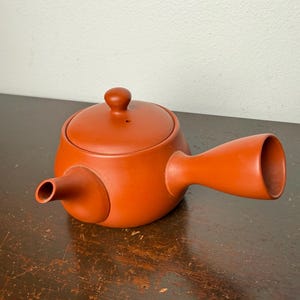 May include: A reddish-brown ceramic teapot with a rounded body, a lid with a small knob, and a side handle. The spout extends from the side of the pot. The teapot is sitting on a dark wooden surface.