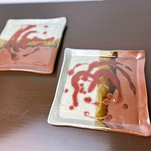 May include: Two rectangular ceramic plates with a white glaze and red and brown abstract designs. The plates are on a brown wooden surface.