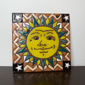 Puede incluir: Azulejo de cerámica cuadrado con un sol sonriente con rayos amarillos, verdes y naranjas. El azulejo tiene un fondo marrón con patrones geométricos y acentos de estrellas en las esquinas. Una pieza decorativa para la decoración del hogar.