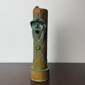 May include: A cylindrical bamboo-style sculpture featuring a carved face wearing a headdress with animal ears. The figure has an open mouth, dark eyes, and is adorned with a necklace featuring leaf-shaped pendants. The surface has green and brown tones.