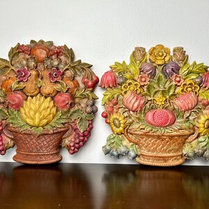 Könnte beinhalten: Zwei Vintage-Wanddekorationen mit 3D-geformten Körben, die überquellend mit Obst und Blumen gefüllt sind. Die Körbe sind in Braun-, Grün- und Gelbtönen bemalt, während die Früchte und Blumen in leuchtenden Rot-, Rosa-, Gelb- und Lilatönen bemalt sind.