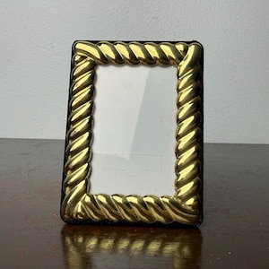 May include: A rectangular picture frame with a gold-colored, rope-like border. The frame has a black backing and a clear glass or plastic insert to hold a photo. The frame is sitting on a dark brown surface.