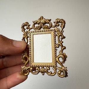 Puede incluir: Un pequeño marco de fotos rectangular de color dorado con un borde ornamentado de volutas, detalles intrincados en forma de hojas y un borde interior con cuentas que rodea un inserto central rectangular blanco para una pequeña fotografía.