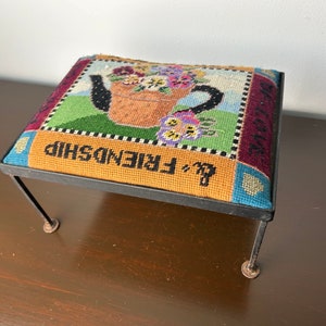 Könnte beinhalten: Ein kleiner schwarzer Metallhocker mit einem bunten bestickten Kissen. Das Kissen zeigt ein Blumenmuster mit einer Teekanne und den Worten "&'s Friendship".