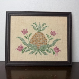 May include: Framed cross-stitch artwork featuring a pineapple design. The pineapple is in shades of brown and gold, surrounded by green leaves and pink flowers. The artwork is set against a beige background and framed in a black wooden frame.