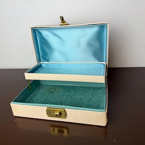 Peut inclure: Une boîte à bijoux rectangulaire de couleur crème avec un couvercle à charnière et un fermoir en métal doré. L'intérieur comprend un plateau supérieur amovible doublé de satin bleu clair et un compartiment inférieur en velours bleu sarcelle.
