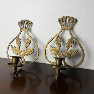 Puede incluir: Dos apliques de pared de latón con un diseño decorativo de hojas. Cada aplique presenta un portavelas y un marco calado detallado. Los apliques están montados sobre una superficie de madera oscura contra una pared blanca.