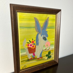K&ouml;nnte beinhalten: Ein gerahmtes Kunstwerk mit einem blauen Esel, der einen Korb mit Blumen tr&auml;gt. Der Esel ist im Cartoon-Stil mit gro&szlig;en Augen und einer rosa Nase dargestellt. Der Hintergrund ist ein gelbes und gr&uuml;nes abstraktes Design. Das Kunstwerk ist unten rechts mit "Dimov 1976" signiert.