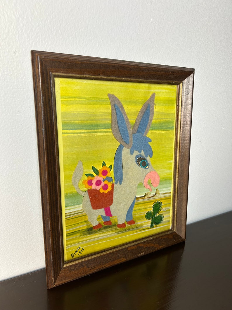 K&ouml;nnte beinhalten: Ein gerahmtes Kunstwerk mit einem blauen Esel, der einen Korb mit Blumen tr&auml;gt. Der Esel ist im Cartoon-Stil mit gro&szlig;en Augen und einer rosa Nase dargestellt. Der Hintergrund ist ein gelbes und gr&uuml;nes abstraktes Design. Das Kunstwerk ist unten rechts mit "Dimov 1976" signiert.