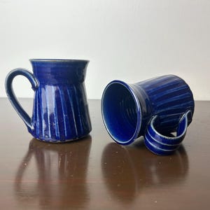 以下が含まれることがあります： ストライプ模様の青いセラミック製のマグカップが2つ。マグカップは濃い茶色の木製の表面の上に置かれています。