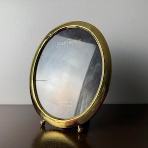 Pode incluir: Moldura oval de latão dourado da coleção Solid Brass. A moldura é polida à mão e mede 12,7 x 17,8 cm. Possui um suporte com três pequenos pés esféricos. A moldura está vazia, com uma inserção de vidro transparente.