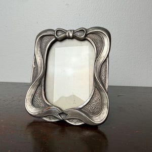 Puede incluir: Un marco de fotos plateado con un lazo en la parte superior y un diseño decorativo y fluido. El marco tiene una abertura rectangular para una foto y se muestra sobre una superficie de madera oscura.