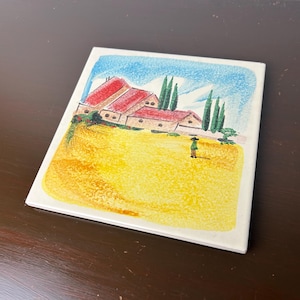 Può includere: Una piastrella di ceramica dipinta a mano che presenta un'illustrazione fantasiosa di un piccolo villaggio con case dai tetti rossi, alberi verdi e una singola figura in un campo. La piastrella ha uno sfondo giallo e bianco.