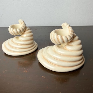 May include: Two off-white ceramic candle holders. Each holder has a spiral base with a curled top, creating a unique, sculptural design. The holders are placed on a dark brown surface, showcasing their creamy color and textured details.