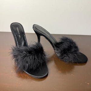 May include: A pair of black high-heeled mules with a fluffy feather detail on the toe strap. The heels are stiletto style. The shoes are on a brown surface. The brand name "VICTORIA'S SECRET" is printed on the insole.