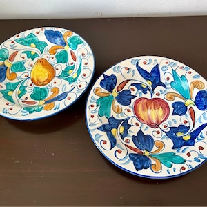 Puede incluir: Dos platos de cerámica blanca con diseños florales azules, verdes, amarillos y rojos. Un plato presenta una pera y el otro una manzana.