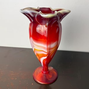 May include: A vibrant red glass vase with a ruffled rim. The vase features a swirling pattern of white, orange, and red. The base is round and the vase is approximately 20 cm tall.