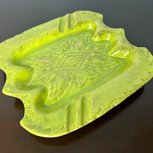 May include: A lime green ashtray with a textured surface and a leaf design in the centre. The ashtray has a unique shape with curved edges and three cigarette rests. A decorative piece for a home.