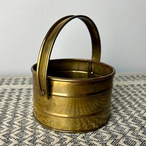 Puede incluir: Un pequeño cubo de latón dorado con asa. El cubo tiene un acabado martillado y es perfecto para guardar objetos pequeños.