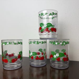 Könnte beinhalten: Set mit vier transparenten Trinkgläsern mit Erdbeermuster. Die Gläser zeigen rote Erdbeeren, grüne Blätter und weiße Blüten. Das Design wiederholt sich am oberen Rand jedes Glases.