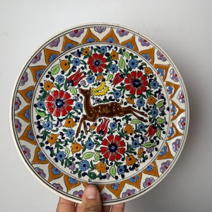 Puede incluir: Plato de cerámica decorativo con un ciervo marrón saltando sobre un diseño floral. El plato tiene un borde blanco con un borde estampado en naranja, amarillo y morado. El diseño floral incluye elementos rojos, azules y verdes.