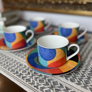 May include: Four matching teacups and saucers. Each cup and saucer features a white interior and a colorful geometric design with blue, yellow, red, and green shapes. The cups have white handles and sit on a patterned tablecloth.