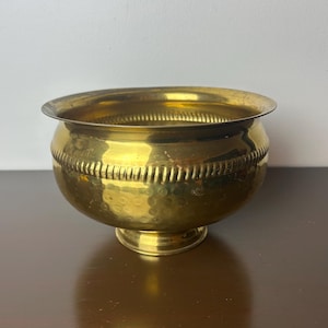 May include: A gold-colored metal planter with a hammered finish and a decorative band around the rim. The planter has a round base.
