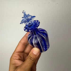 May include: A small cobalt blue glass perfume bottle featuring vertical white stripes and a decorative glass stopper shaped like a flower. The bottle has a rounded body and a narrow neck, with a translucent finish that catches the light.