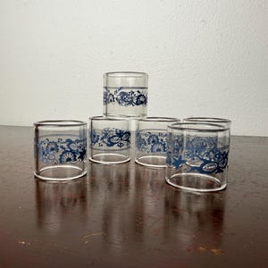 Puede incluir: Seis vasos de vidrio transparente con un diseño decorativo floral y de volutas azul en la parte superior. Los vasos están dispuestos en forma de pirámide sobre una superficie de madera oscura, mostrando su elegante diseño.