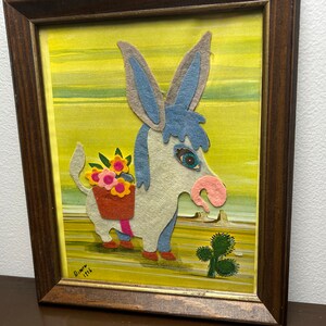 K&ouml;nnte beinhalten: Ein gerahmtes Filz-Applikationsbild, das einen blauen und grauen Esel zeigt, der einen Korb mit Blumen tr&auml;gt. Der Esel steht auf einem gelben Hintergrund mit einer gr&uuml;nen Pflanze im Vordergrund. Das Kunstwerk ist signiert mit "Diann 1976".