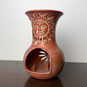 May include: A terracotta-colored ceramic oil burner featuring a sculpted sun face on the upper neck and a large circular opening at the base. The design includes cut-out sun rays around the face and within the lower chamber, with a matte finish.