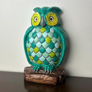 May include: A teal owl figurine with yellow eyes and accents. The owl is perched on a brown log base. The body is decorated with a scale pattern in shades of teal, green, and yellow.