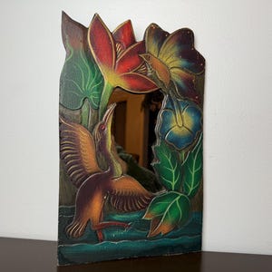 May include: Rectangular wooden mirror with a hand-painted design of a bird, flowers, and leaves. The bird is brown and orange, with a long beak and outstretched wings. The flowers are red, blue, and yellow, and the leaves are green.