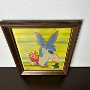 K&ouml;nnte beinhalten: Ein gerahmtes Kunstwerk mit einem blauen und grauen Esel mit rosa Nase, der einen Korb mit Blumen tr&auml;gt. Der Esel ist vor einem gelben Hintergrund mit gr&uuml;nen Streifen platziert. Der Rahmen ist aus braunem Holz.