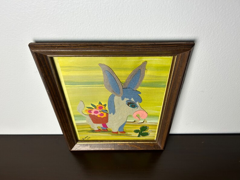 K&ouml;nnte beinhalten: Ein gerahmtes Kunstwerk mit einem blauen und grauen Esel mit rosa Nase, der einen Korb mit Blumen tr&auml;gt. Der Esel ist vor einem gelben Hintergrund mit gr&uuml;nen Streifen platziert. Der Rahmen ist aus braunem Holz.