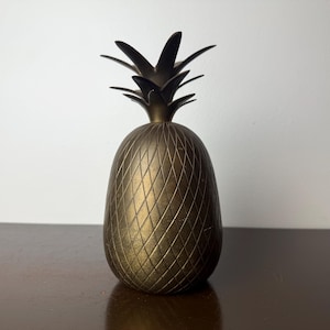 May include: A gold-colored pineapple-shaped decorative object. The pineapple has a textured surface with a diamond pattern. The top of the pineapple features a crown of spiky leaves.