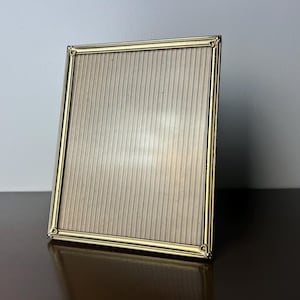 May include: A rectangular gold-colored picture frame with a decorative border. The frame is empty, with a light beige, vertically-striped backing. The frame is angled slightly, resting on a dark brown surface, with a light gray background.